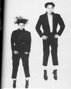 Two dancers in black pants, jackets, and shoes, jumping in the air at different heights