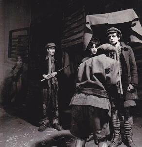 Four actors in tattered clothes speaking, with an actor dressed as a German soldier in the background