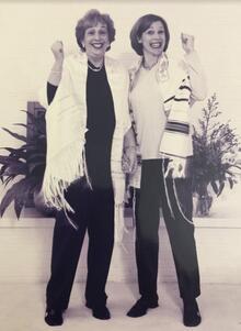 Two women smiling at the camera, gesturing happily and wearing prayer shawls