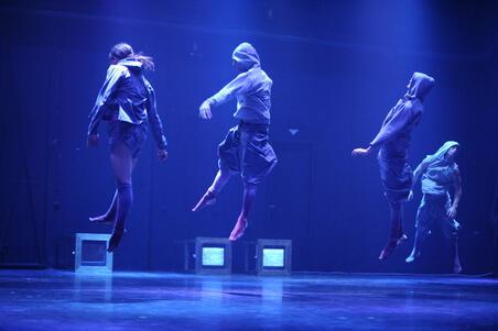 Four dancers in hooded sweatshirts leaping in the air, with the stage lit blue