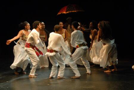 A group of dancers in a circle on stage, with the men wearing loose white trousers and long sleeved shirts with red patterned cloth belts, and the women long white skirts and patterned sleeveless tops