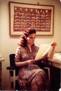 Ruth Behar sitting in a chair beside a desk, reading a piece of paper, beneath a large framed tapestry hanging on the wall