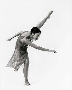 Ze'eva Cohen, dressed in a skirted leotard, striking a dance post with one foot on the ground and the other lifted above her head
