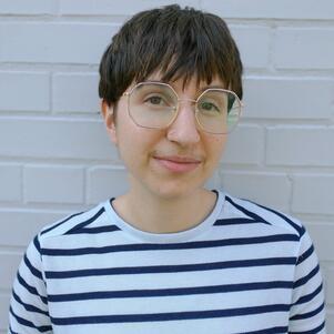 Young person with short brown hair and wire-rimmed glasses wearing a white t shirt with black stripes, posing against a white brick wall