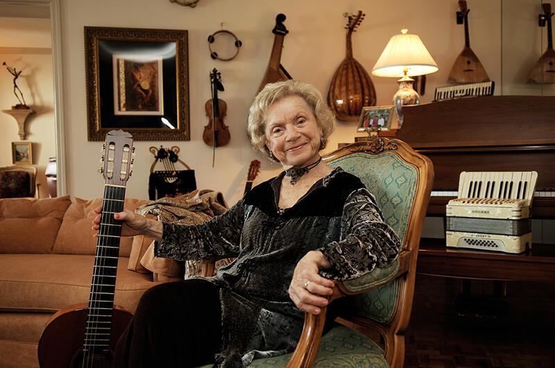 Flory Jagoda sitting in a chair, holding the neck of a guitar, with more instruments hanging on the wall behind her