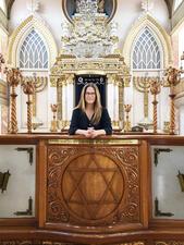 Mónica Unikel-Fasja standing before the Torah ark in an elaborately decorated synagogue with carved candelabras, menorahs, and columns.