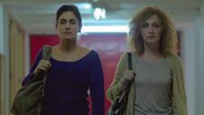 A woman with dark hair in a bun in a blue top, left, and a woman with curly blonde hair stand side in fluorescent lit space. Both have right arm holding purses in place on shoulder. 