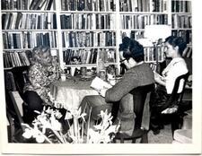 phtoograph of a woman in a flowered shirt sitting at a table with a man and a woman, looking intently at them, in front of a wall of bookshelves