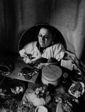 black and white photograph of the reflection in a mirror of a woman wearing glasses and a white blouse, looking at a piece of jewelry she is making. Supplies for making jewelry are on a table in front of the mirror.