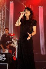 Photograph of a woman with dark hair wearing a black sparkly dress singing at a microphone, with a man playing drums in the background