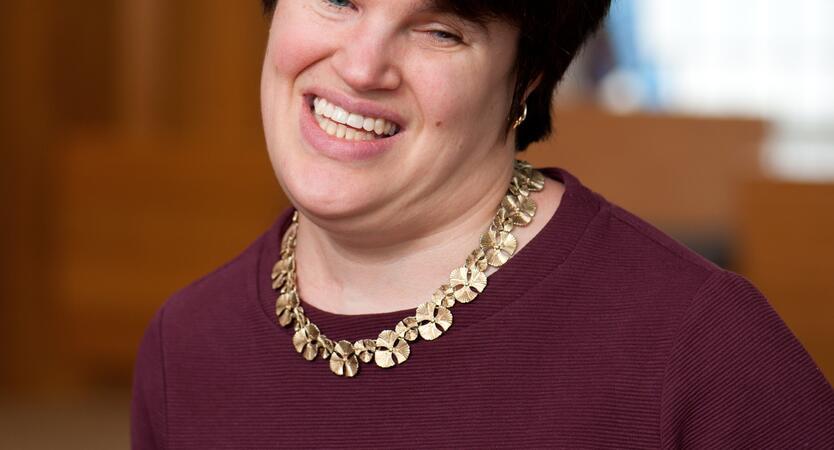 Portrait of Lauren Tuchman smiling in front of a stained glass window wearing a maroon top and gold necklace