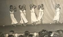 A group of six women, barefoot, wearing long white tunics, and holding up clay pots as they dance onstage