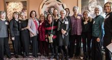Group of 13 women smiling and posed, standing  for group photo in front of art