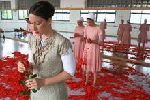 A dancer in a patterned dress holding a rose in a dance studio with a mirrored wall. Behind her stand four other dancers in pink dresses and white headscarves, holding red rose petals, which are scattered heavily over the floor.