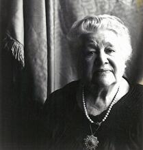 Black and white phtoograph of a woman with white hair wearing a dark dress looking pensively at the camera
