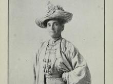 Black and white historical photo of a woman wearing a light colored dress and a large hat, with a serious expression