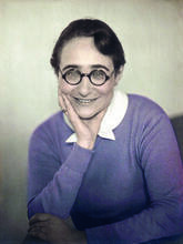 photograph of a seated woman wearing a purple sweater and round glasses, with her chin in her hand, smiling at the camera