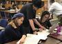 Several small groups of women studying at small tables, with teachers leaning over to assist them