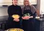 Two women standing in a kitchen, one holding a strainer with noodles and the other a strainer with greens