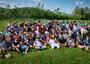 Large group of people sitting on grass, hills in background. 