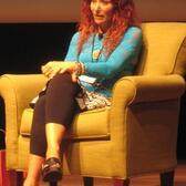 Aline Kominsky speaking on stage, sitting in an armchair