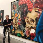 Audrey Flack wearing acid wash jeans and a black top standing in front of a large painting with many items including a skull, tarot, lipstick, candle