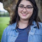 Photo of Lila Goldstein, in front of a tree trunk wearing a striped blue and white shirt and a denim overshirt.