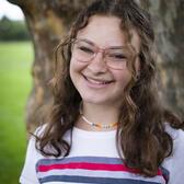 Photo of Neima Fax, in front of a tree wearing a white shirt featuring a multicolor horizontal stripe.