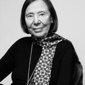 Black and white photo of Grace Schulman, seated in a black outfit with a scarf and locket or pocket watch necklace