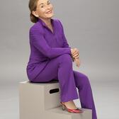 Judy Sheindlin smiling to camera while sitting on grey prop stairs in purple suit and heels in front of grey backdrop