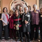 Group of 13 women smiling and posed, standing  for group photo in front of art