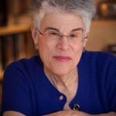 Judith Hauptman wearing a blue top and glasses smiling into the camera with arms crossed over one another