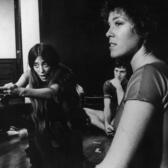 Liz Swados kneeling on a stage with both arms stretched forward, directing four performers who sit in a half-circle around her