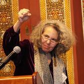 Writer and editor Melanie Kaye Kantrowitz standing at a podium making a clenched fist of solidarity