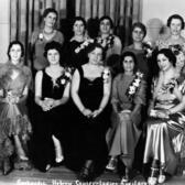 5 women seated in the front row and 5 women standing in the back row, smiling and wearing formal dresses
