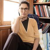 Joey Soloway wearing dark pants, yellow top, and glasses, seated in front of a window and a bookcase with right arm propped up on bent right leg