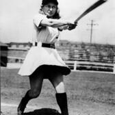 Thelma Eisen swinging a bat, wearing a skirted baseball uniform and a hat reading "GR"