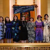 A group of seven women standing at a bimah, posing underneath a banner with Hebrew writing