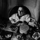 black and white photograph of the reflection in a mirror of a woman wearing glasses and a white blouse, looking at a piece of jewelry she is making. Supplies for making jewelry are on a table in front of the mirror.