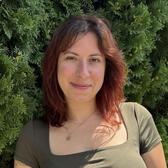 Emma Breitman stands smiling in front of green foliage. She has reddish brown hair and is wearing an olive green shirt.