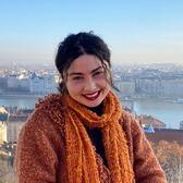 Aliza smiles at the camera. She is wearing an orange scarf and has a cityscape behind her.