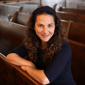 Rabbi Lila Kagedan sits on a pew leaning forward with her arms crossed and resting on the back of the pew in front of her