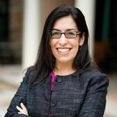 Lisa Lepson, smiling, stands in front of a blurred background of white pillars. Lisa has long dark hair, glasses, and is wearing a grey jacket and pink shirt.