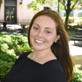 Madisen Siegel is a young woman with reddish brown hair, light freckles, and blue eyes. She is wearing a black top and is smiling against a backdrop of plants. 