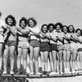 Thirteen young women stand, smiling and laughing, and pose with hands on each others' shoulders for a photo