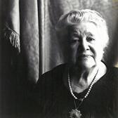 Black and white phtoograph of a woman with white hair wearing a dark dress looking pensively at the camera
