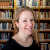 Headshot of Rachel Barenbaum. She is a light skinned woman with blonde hair and blue eyets. She is smiling and wearing a black top in front of a wall of bookshelves.