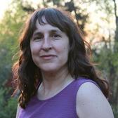 Headshot of Rachel Kranson. She is a light skinned woman with wavy dark brown hair. She is wearing a purple tank top, with a backdrop of sky and trees. 