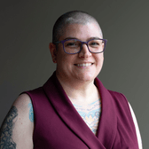 Wendy has buzzed hair, purple glasses, and several tattoos. She is wearing a red v-neck shirt and is in front of a gray background.