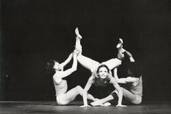 Miguel Angel Palmeros and Bernardo Benitez sitting on a stage, lifting Cecilia Baram's legs in the air as she places her palms on the floor and looks up at the audience